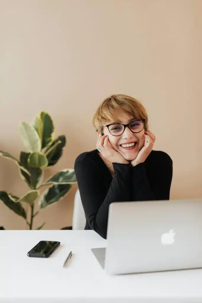 A smiling lady wearing glasses, sitting at a white table with a laptop, mobile phone, and pen, with a potted plant and light brown wall in the background.