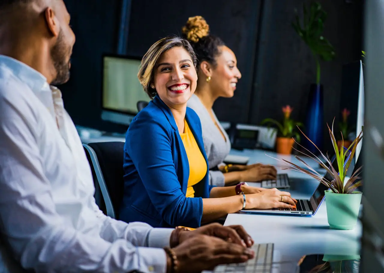 A smiling lady in a yellow T-shirt and blue jacket sitting with two co-workers at their desks, working happily.